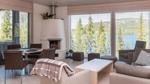 Una sala de estar moderna con chimenea, asientos cómodos y grandes ventanales que ofrecen vista a árboles y un cuerpo de agua.