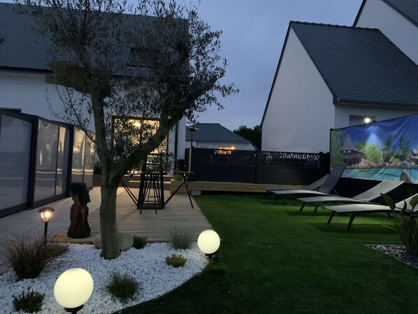 Una casa moderna con un patio, un árbol y un área de piscina con sillones de descanso.