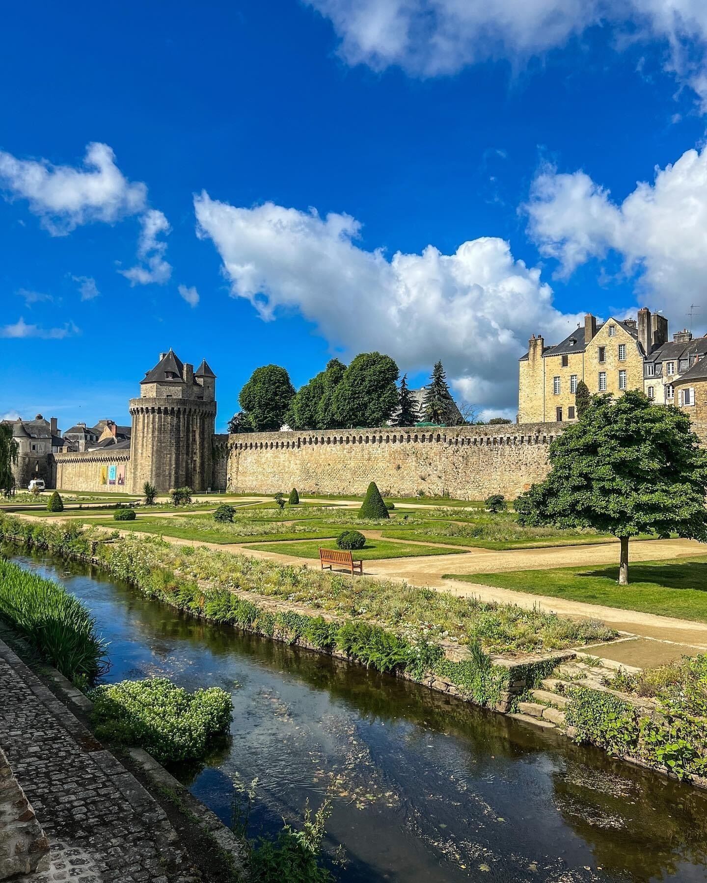 A historic stone wall with a river in the foreground, a bench, and a large building with a tower in the background.