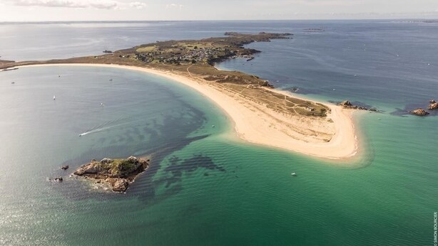 An aerial view of a coastal area with a sandy beach, clear waters, and a small island.
