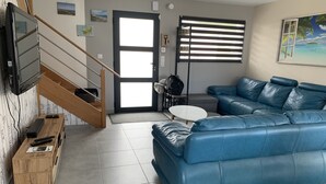 Uma sala de estar moderna com um sofá de couro azul, uma escrivaninha de madeira com uma TV, uma mesa pequena e uma escada que leva a outro cômodo.