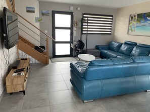 Una sala de estar moderna con un sofá de cuero azul, un escritorio de madera con un televisor, una mesita pequeña y una escalera que lleva a otra habitación.