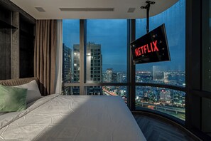 Quarto com uma janela ampla, uma cama e uma TV fixada na parede exibindo o logotipo da Netflix.