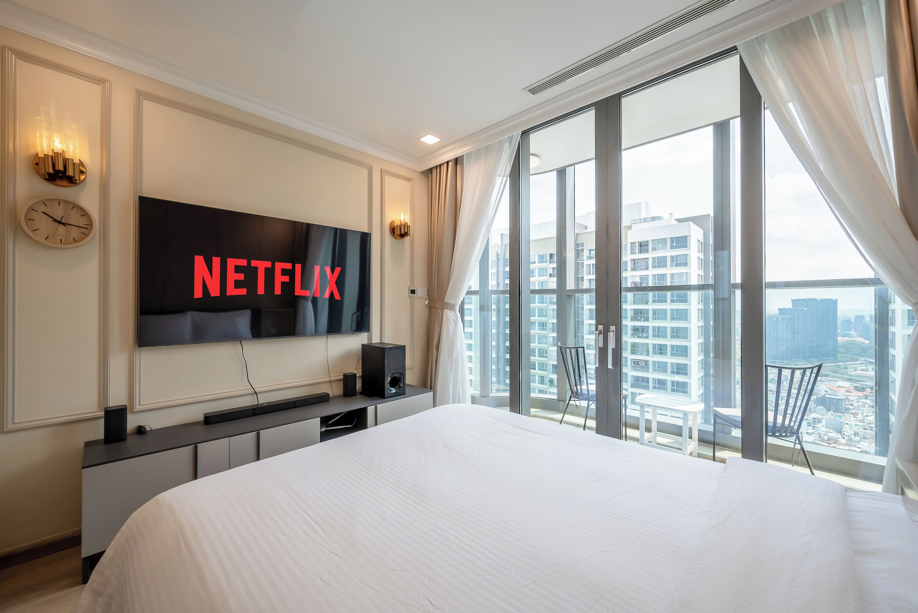 Habitación de hotel con una cama grande, un televisor en la pared mostrando 'NETFLIX', un reloj y vistas de un paisaje urbano a través de ventanas amplias.