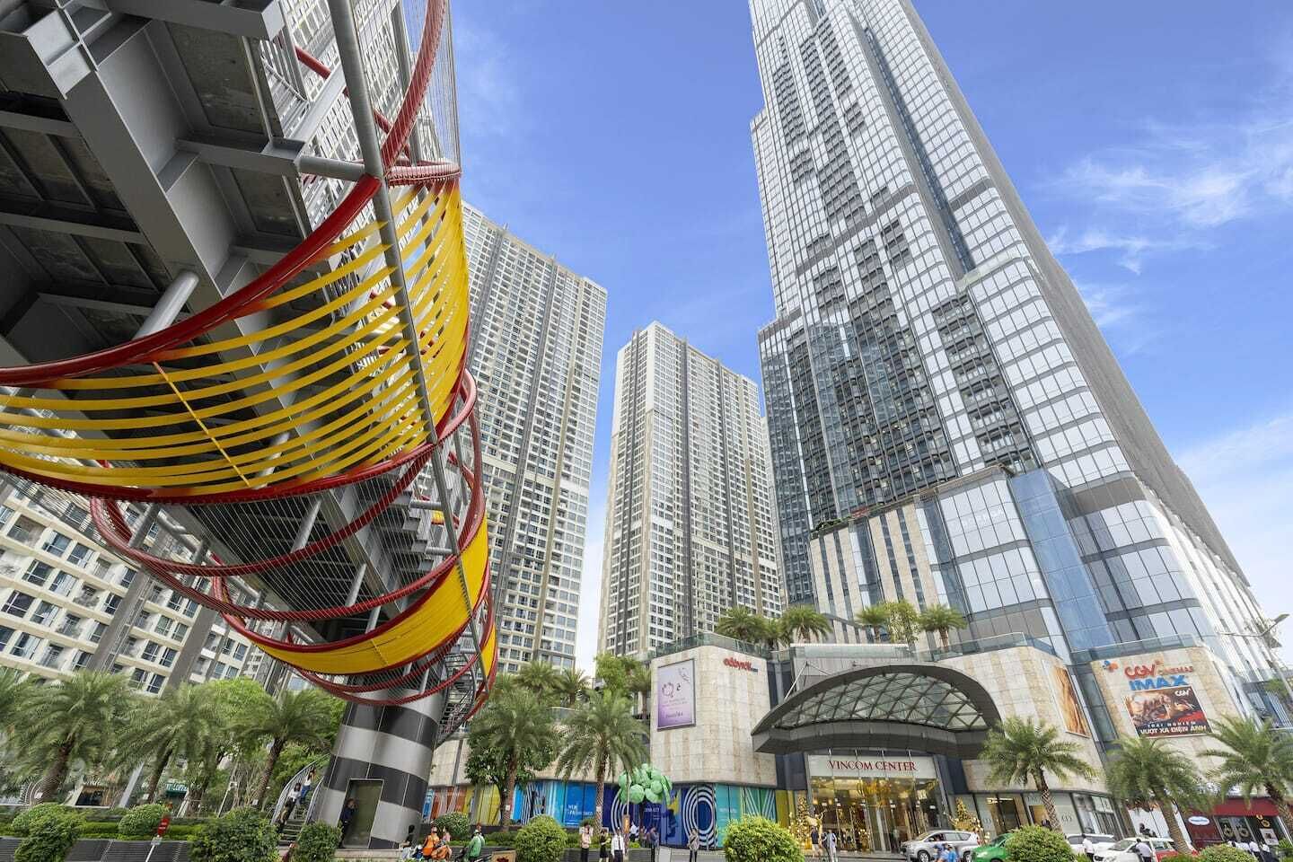 Una zona urbana moderna con edificios altos, una estructura destacada en amarillo y rojo, y el Vincom Center.