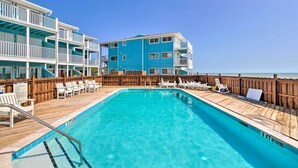 A pool area with a wooden deck, lounge chairs, and a building with balconies. - Kure Beach Condo w/ Panoramic Ocean Views! (Kure Beach)