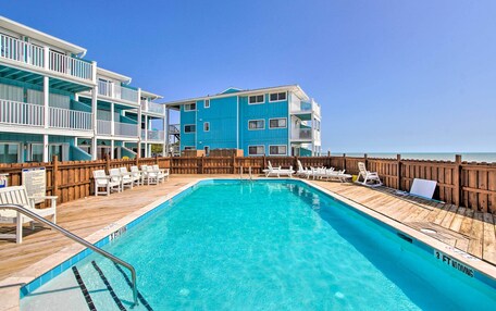 A pool area with a wooden deck, lounge chairs, and a building with balconies.