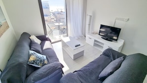 A modern living room with a grey sofa set, a coffee table, a TV on a white stand, and a balcony with a table and chairs. - Sea La Vie Seaview Apartment (Larnaca)