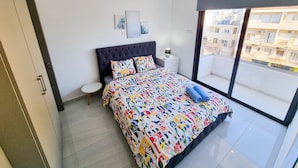 A modern bedroom with a large bed, a nightstand, a window with a view of buildings, and a ceiling fan. - Sea La Vie Seaview Apartment (Larnaca)