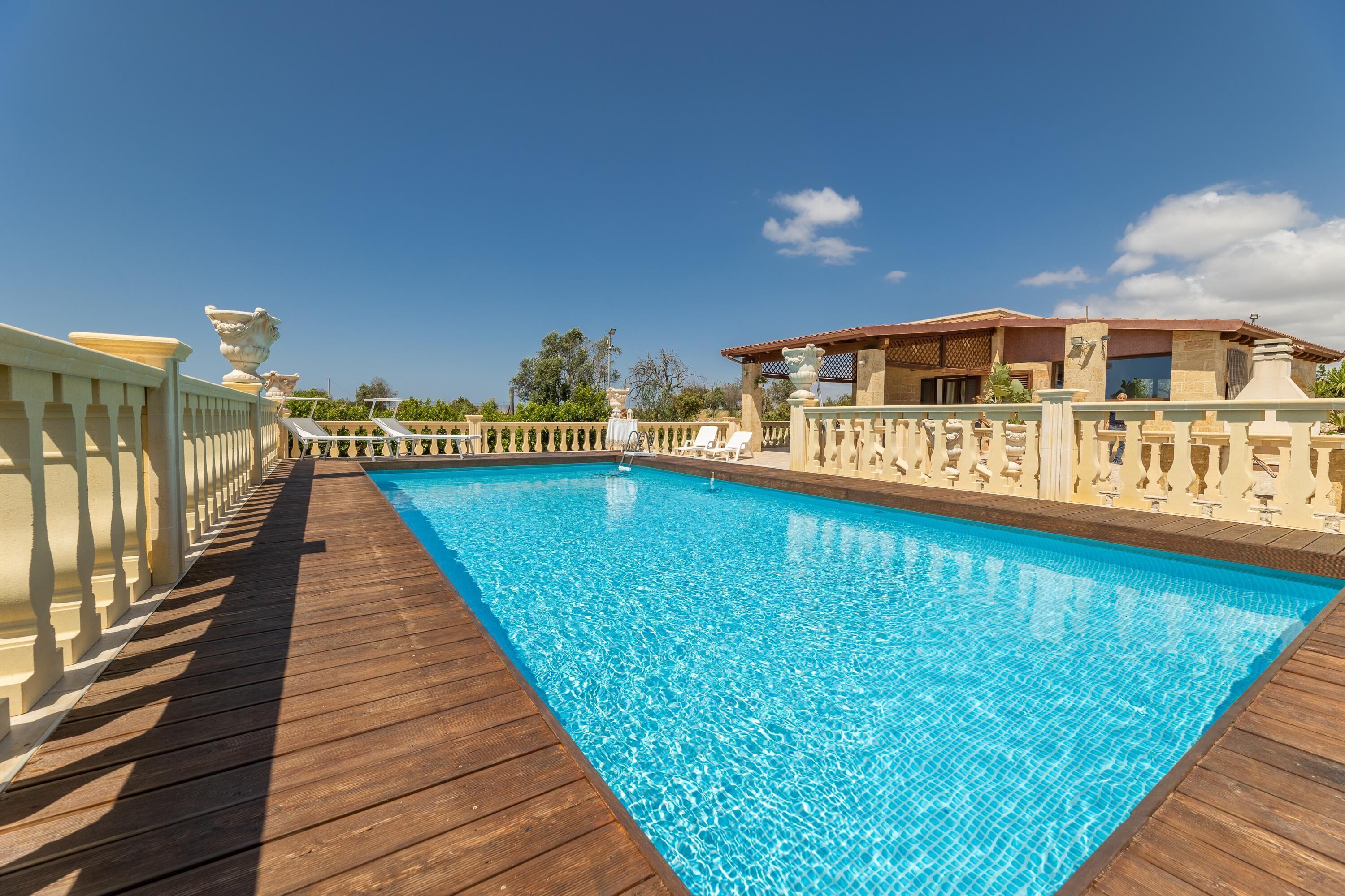 A swimming pool with a wooden deck, a pergola, and a building with a tiled roof.