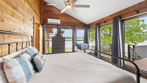 A bedroom with a large bed, wooden walls, a ceiling fan, and a view of the outdoors. - Crystal Springs - Lakefront with Hot Tub! (Linn Creek)
