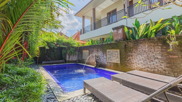 A swimming pool with lounge chairs, surrounded by lush greenery and a brick wall with decorative tiles. - Komala Homestay by ecommerceloka (Canggu)