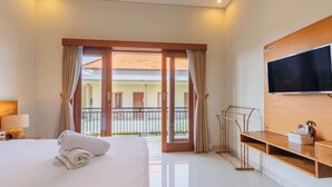 A hotel room with a large bed, a TV mounted on the wall, a wooden desk, and a balcony with a view of the building's exterior. - Komala Homestay by ecommerceloka (Canggu)