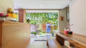 A modern interior with a wooden reception desk, a bench, and a view of a garden with a brick wall and greenery. - Komala Homestay by ecommerceloka (Canggu)
