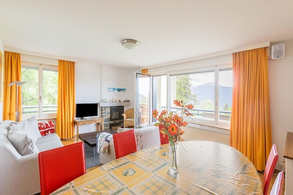 Un salon moderne avec un canapé, une table à manger et un téléviseur. De grandes fenêtres sont agrémentées de rideaux, et on y trouve une cheminée ainsi qu’une vue sur l’extérieur.