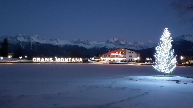 Un hotel con letreros iluminados que dicen "CRANS MONTANA" en un paisaje nevado con montañas al fondo.
