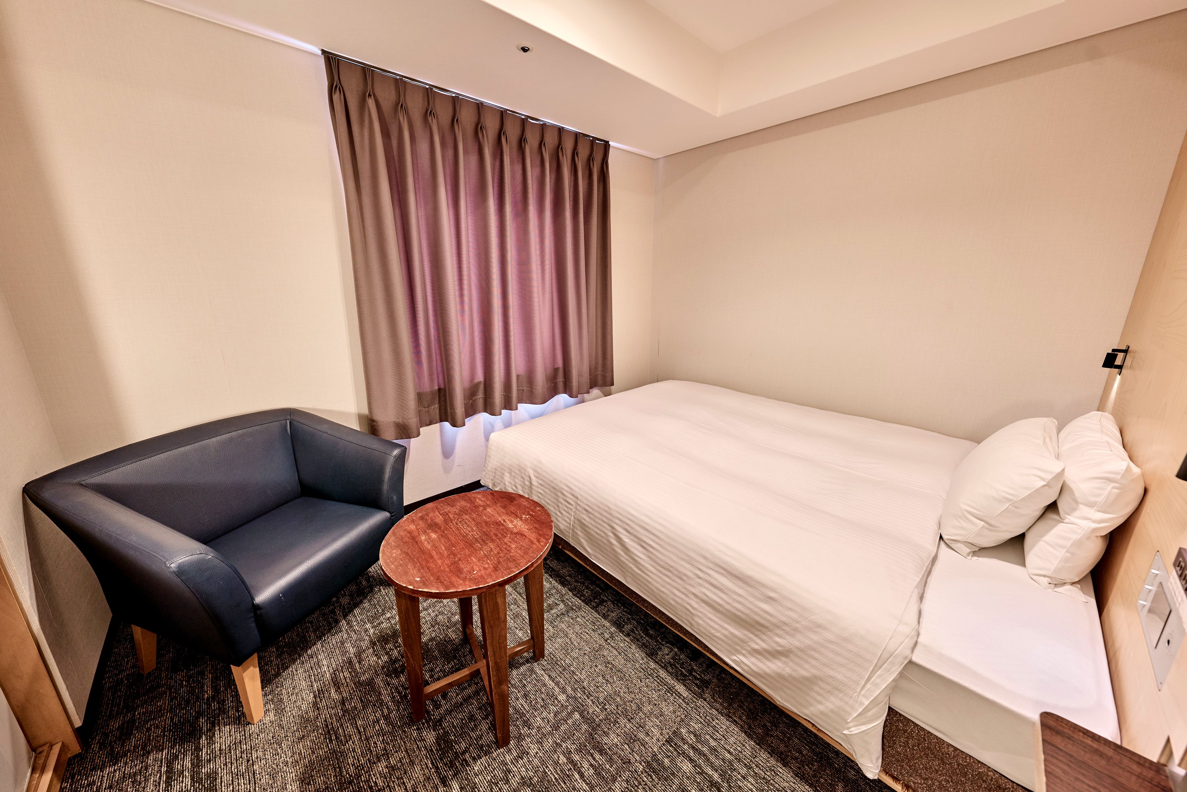 A hotel room with a single bed, a small table, a chair, and a window with curtains.