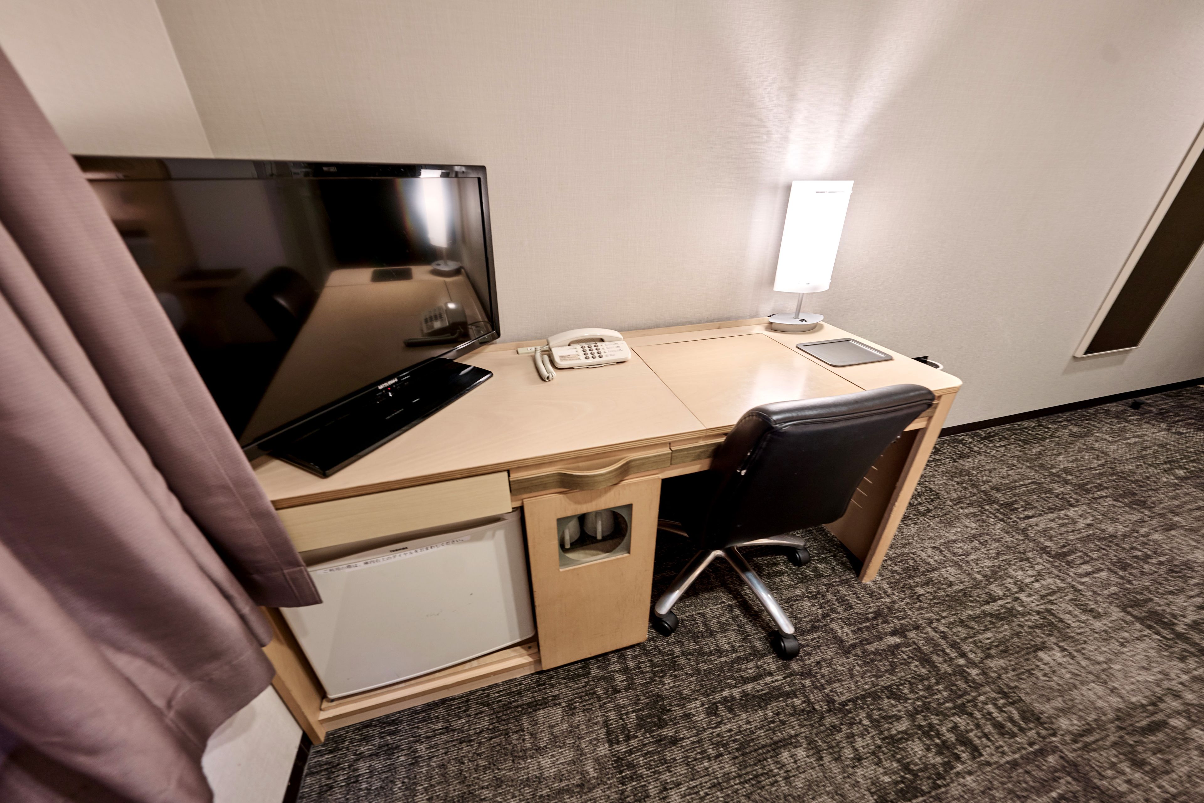 A hotel room with a desk, a computer, a telephone, a lamp, and a small refrigerator.