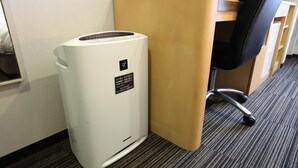 Een witte luchtreiniger met een label, geplaatst naast een houten bureau en een zwarte bureaustoel in een kamer met vloerbedekking.