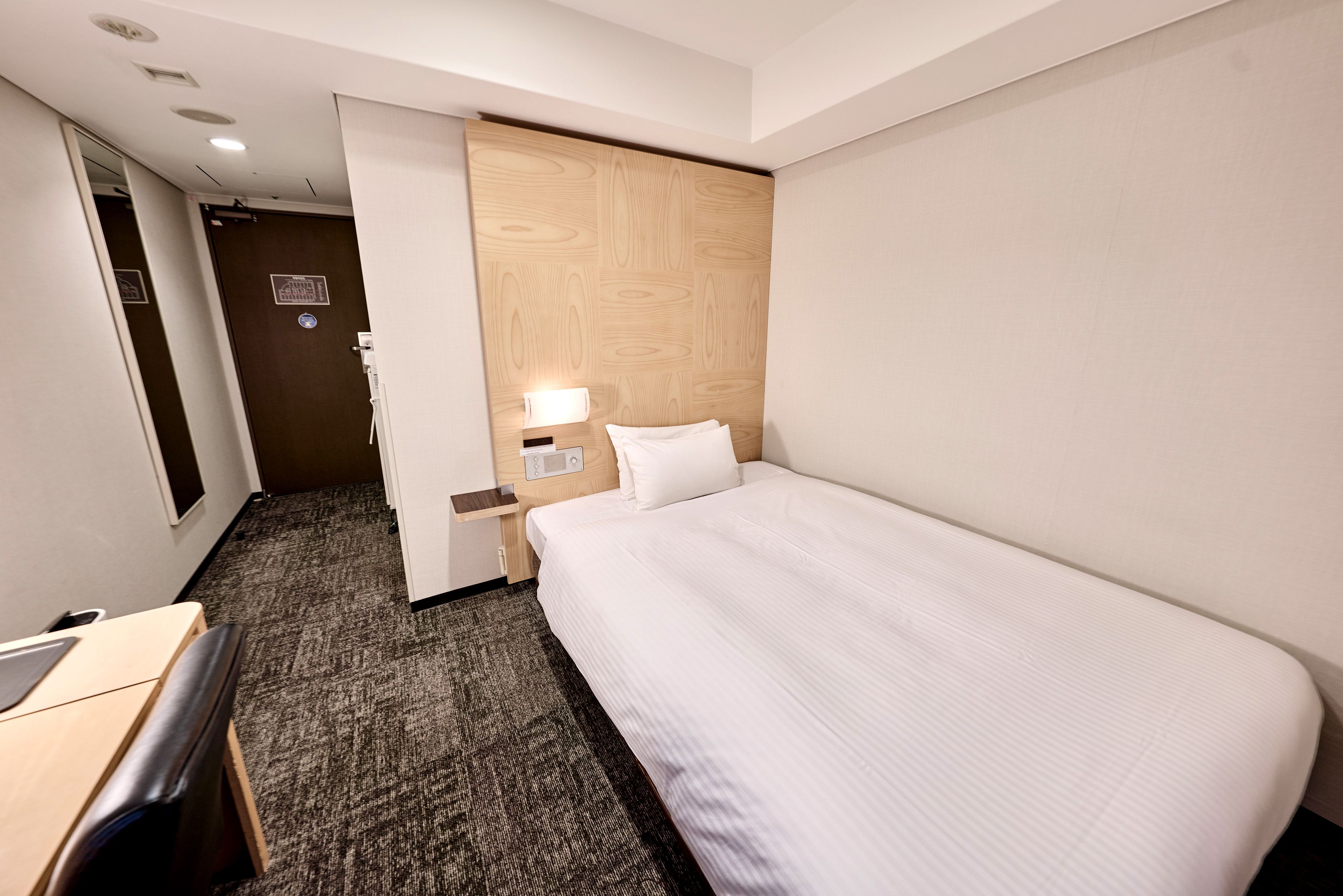 Une chambre d’hôtel avec un lit, un bureau, une chaise, une table de chevet, une lampe et une porte.
