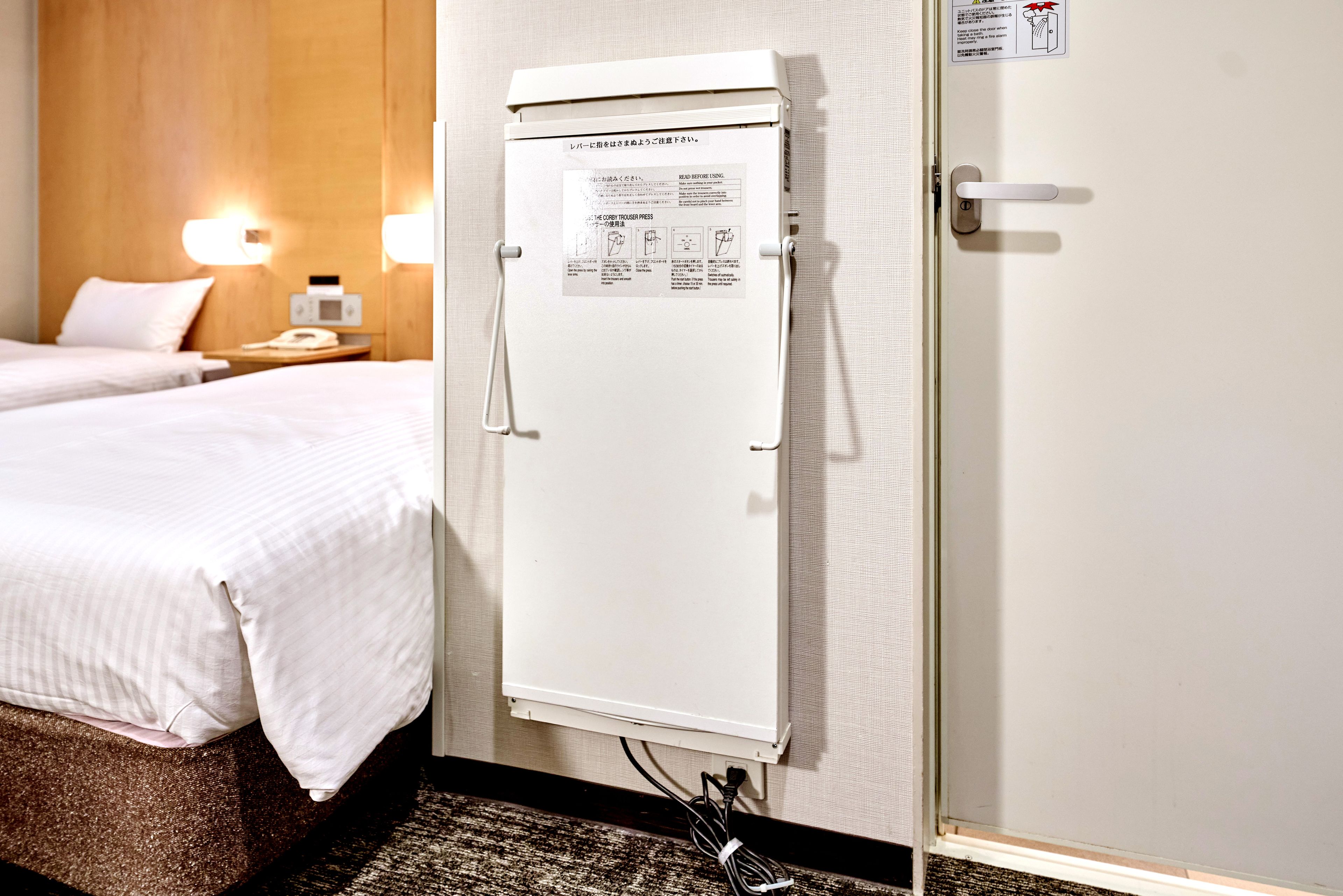 Een hotelkamer met een bed, een nachtkastje, een lamp en een witte, aan de muur bevestigde apparatuur met instructies.