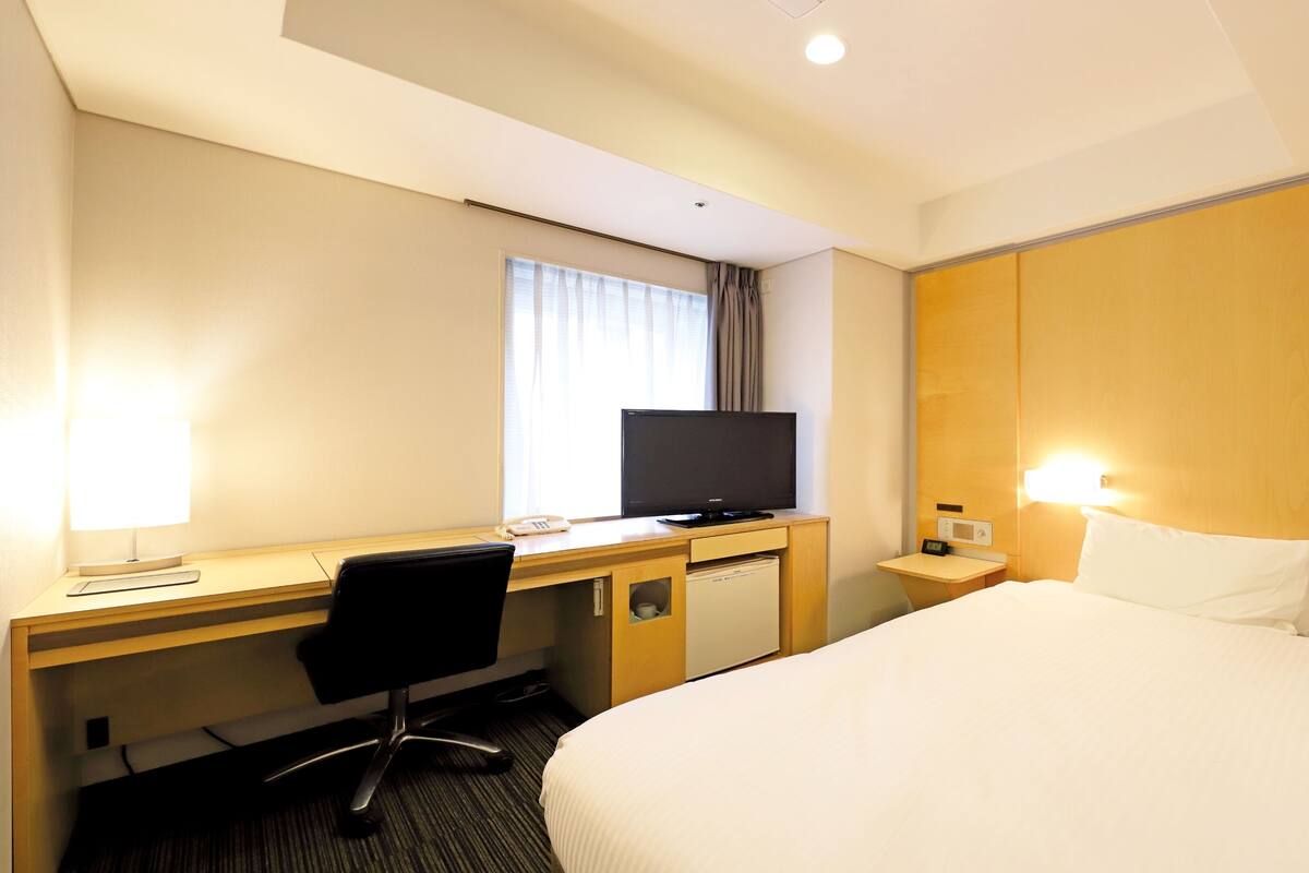 Une chambre d’hôtel comprenant un lit, un bureau avec un ordinateur, une chaise, une télévision, une table de chevet avec une lampe et une fenêtre avec des rideaux.