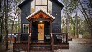 A cabin with a wooden front door, a porch with two chairs, and a large window above the door. - Cozy Modern Farmhouse Hot Tub, Firepit, Game Table (Broken Bow)