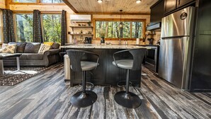 A modern kitchen with a central island, bar stools, a refrigerator, and a dining area with a sofa and coffee table. - Cozy Modern Farmhouse Hot Tub, Firepit, Game Table (Broken Bow)