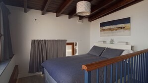 Un dormitorio con cama, cabecero de madera, ventana con cortinas, un cuadro en la pared y una lámpara colgante.