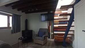 Una habitación con una cama, televisor, sillón, mesita, ventana con cortinas y una escalera con un letrero.