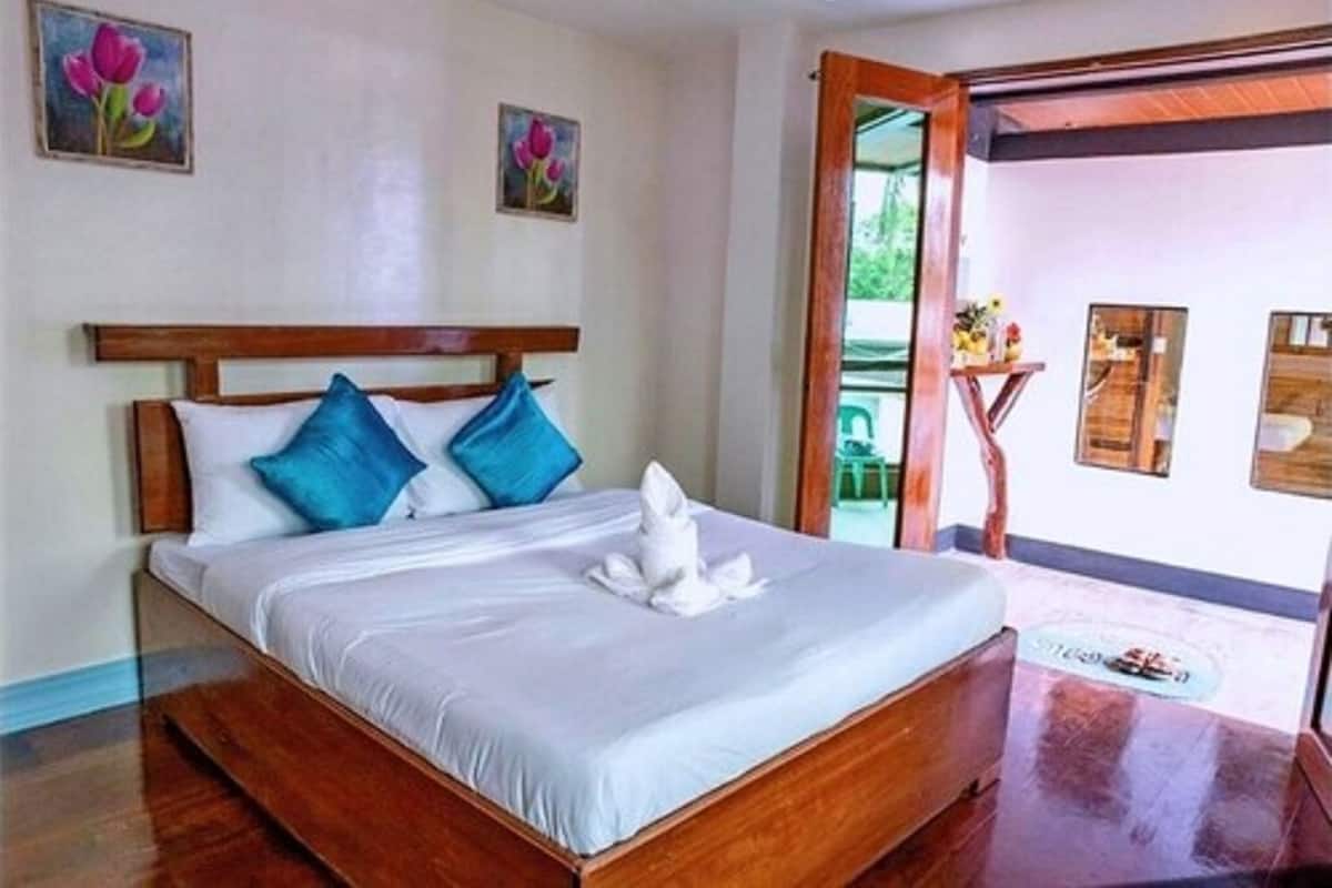 Un lit bien fait, avec des oreillers bleus et blancs, une tête de lit en bois et une serviette blanche posée sur le lit. Deux tableaux ornent le mur et un miroir se trouve sur le côté droit.