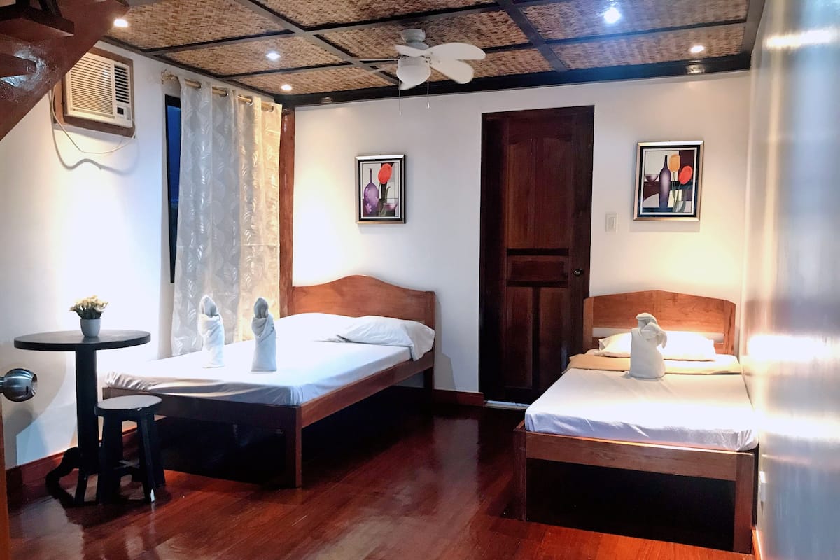 Une chambre d’hôtel avec deux lits, une petite table sur laquelle est posé un vase, et un ventilateur de plafond.