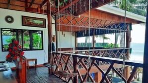 Une terrasse en bois offrant une vue sur l’océan, un coin repas avec une table et des chaises, et une balançoire suspendue.