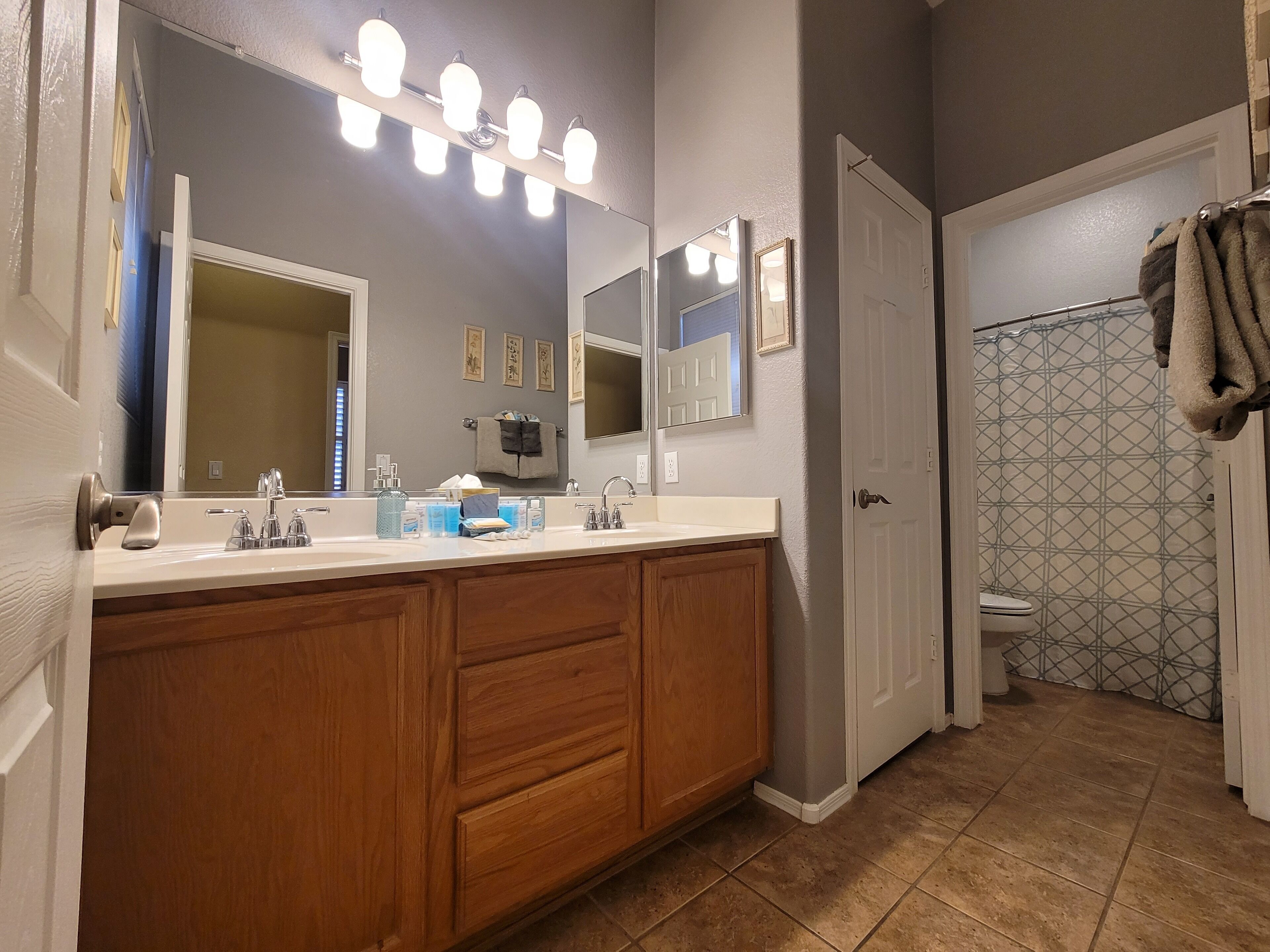Une salle de bain dotée d’un meuble-lavabo à deux vasques, d’armoires en bois, d’un grand miroir avec éclairage intégré, d’une douche avec un mur recouvert de carreaux à motifs et d’un porte-serviettes.