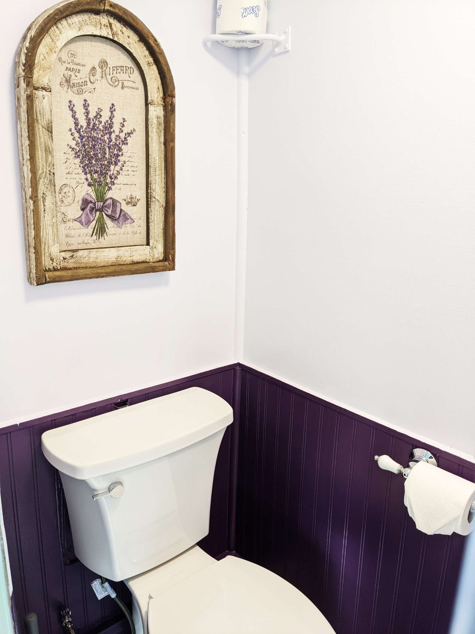 Un baño con inodoro blanco, paneles de pared morados y un cuadro con flores de lavanda en la pared.