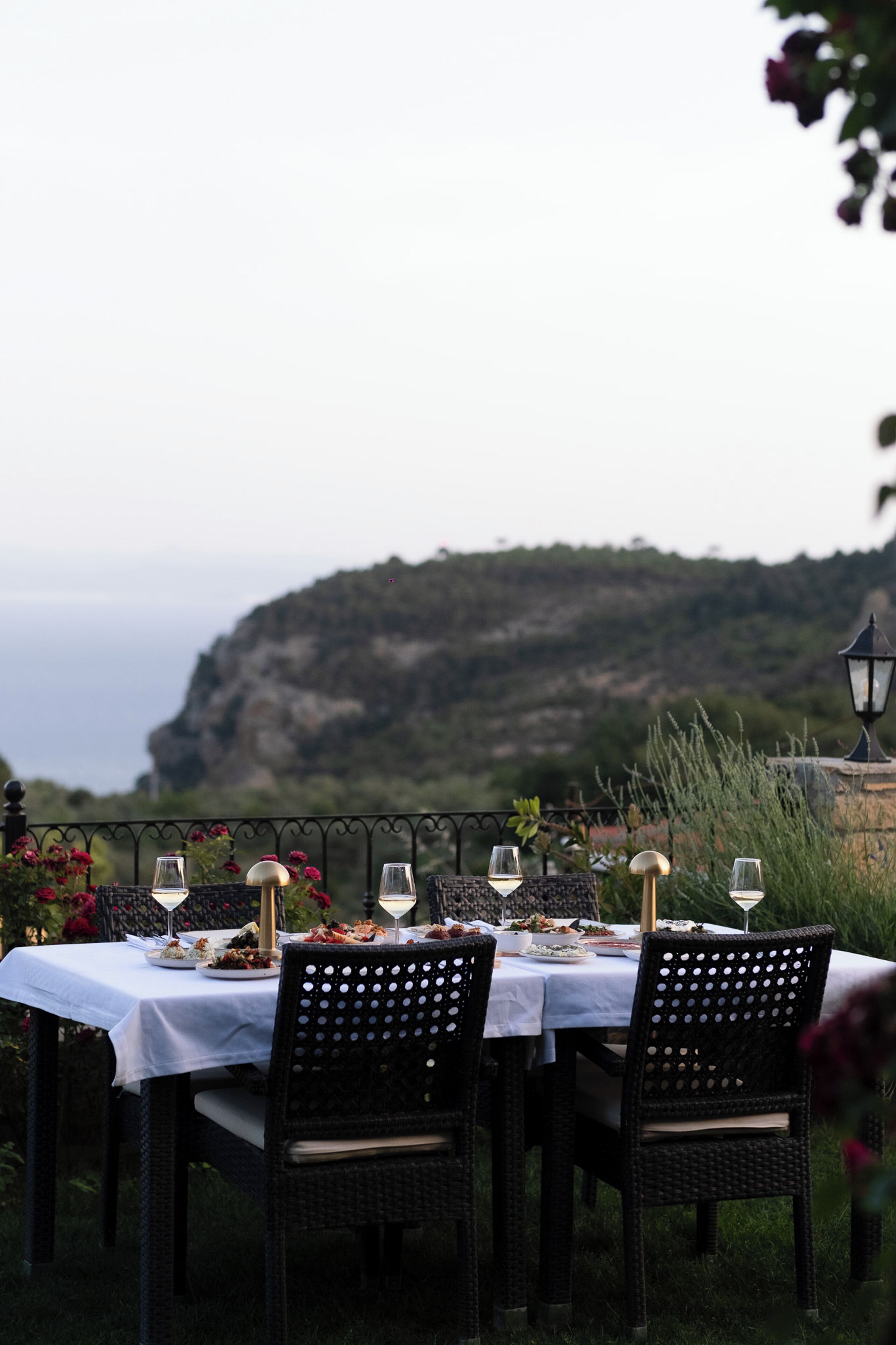 Una mesa de comedor al aire libre con manteles blancos y sillas de mimbre negro, frente a una vista panorámica de un acantilado y el mar.