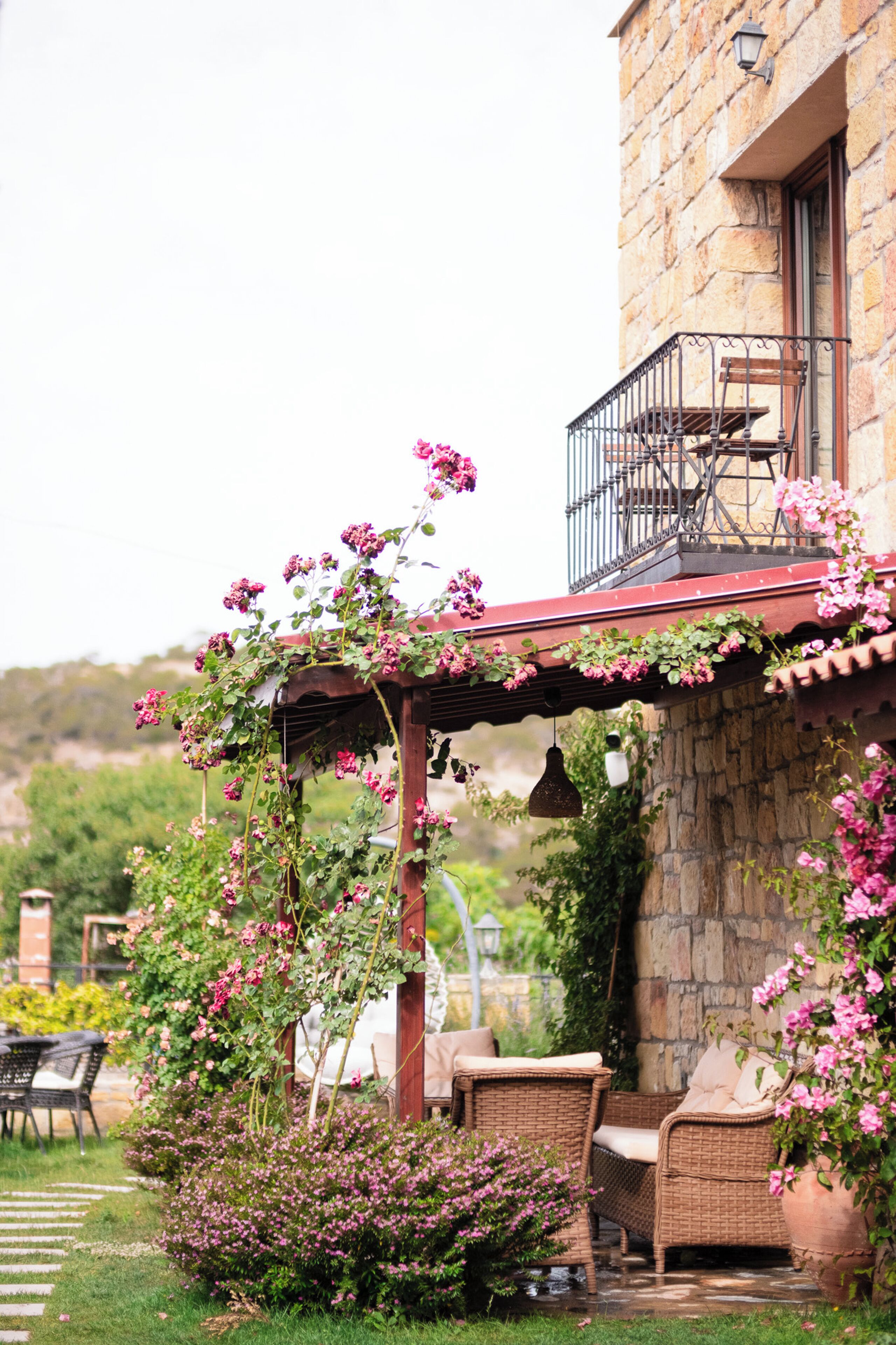 A stone building with a wooden pergola covered in pink flowers, outdoor wicker furniture, and a stone pathway.