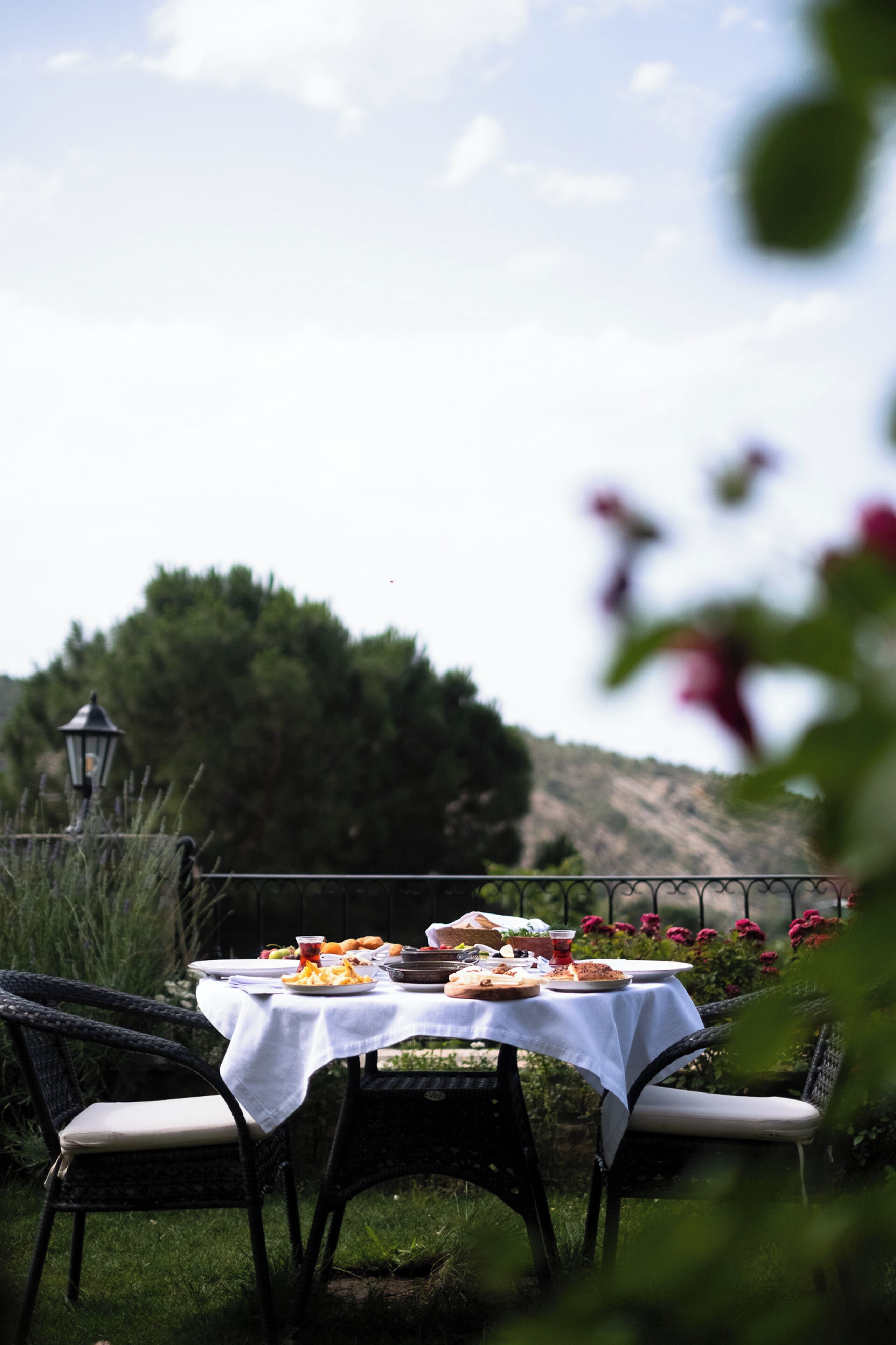 Mesa de comedor al aire libre preparada con platos, vasos y comida, rodeada de sillas de mimbre, con vista a una colina y un farol al fondo.