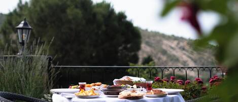 Mesa de comedor al aire libre preparada con platos, vasos y comida, rodeada de sillas de mimbre, con vista a una colina y un farol al fondo.