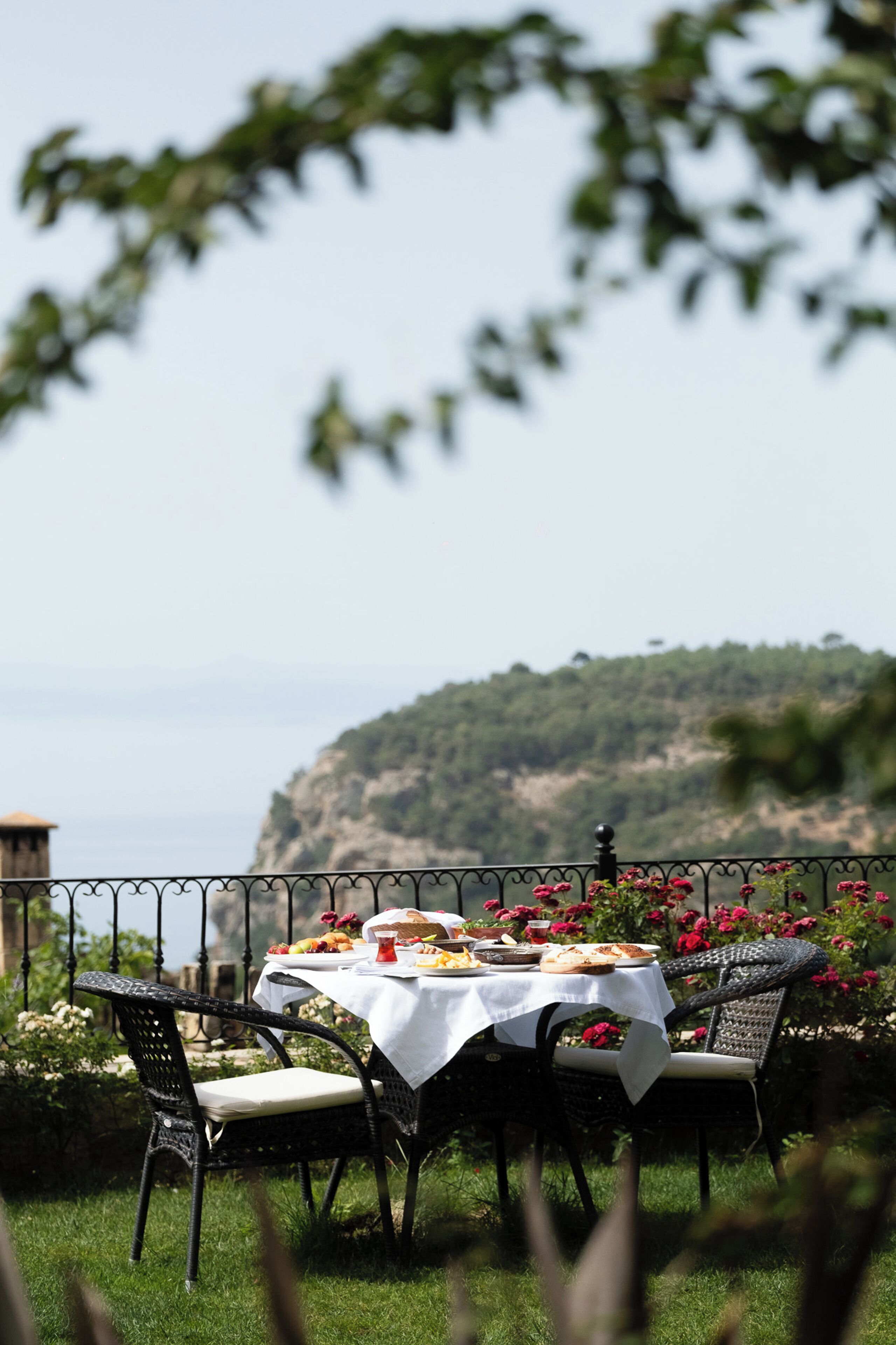 An outdoor dining table with a white tablecloth, surrounded by wicker chairs, set against a scenic hillside backdrop.