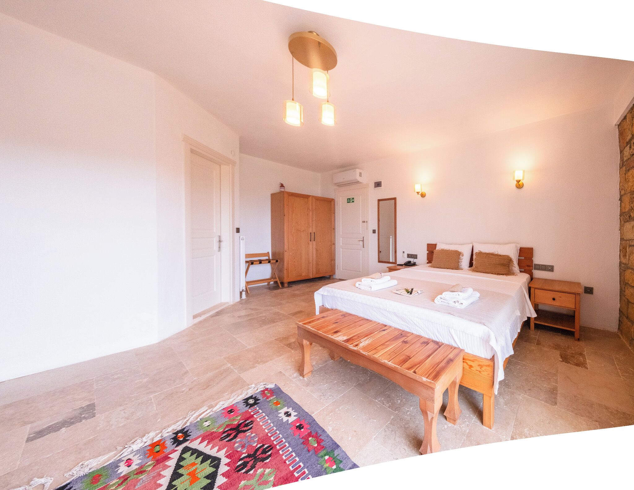 Una habitación de hotel con una cama grande, un banco de madera, un tapete colorido y una pared de piedra.