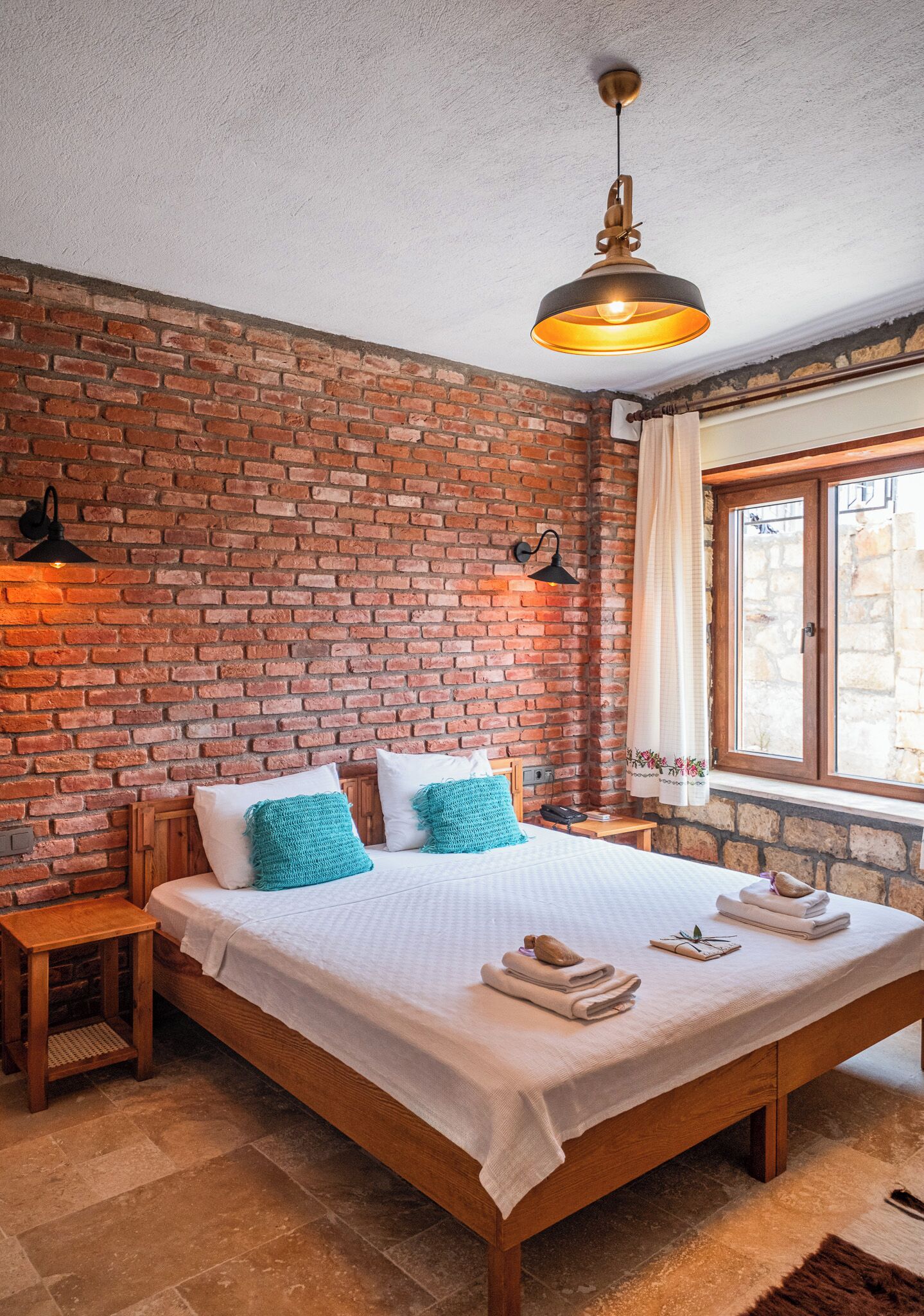 Un dormitorio con pared de ladrillo, una cama de madera, dos mesitas de noche, una lámpara colgante y una ventana con cortinas.