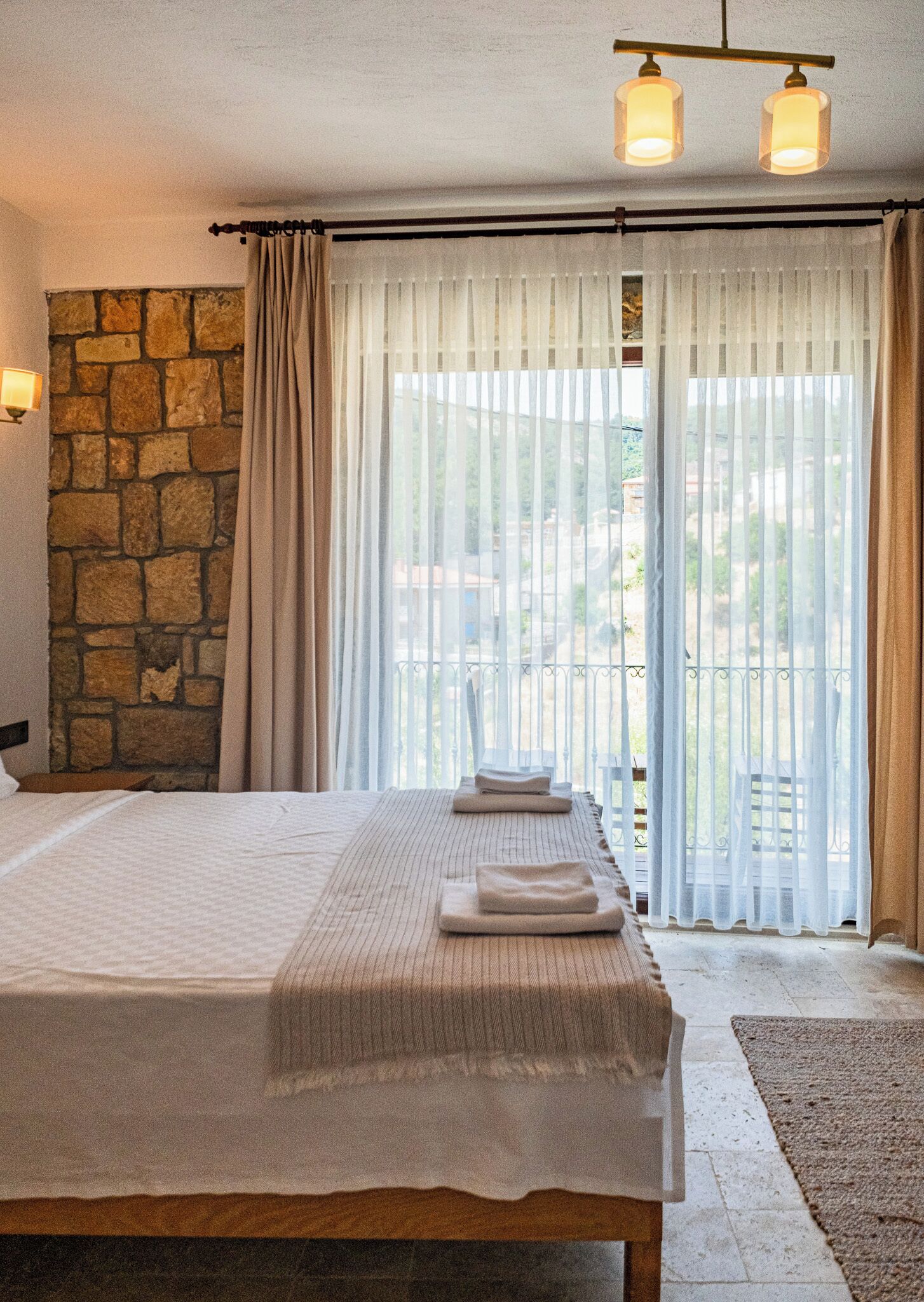 A bedroom with a stone wall, a bed with a white bedspread, two towels, and a balcony with white curtains.