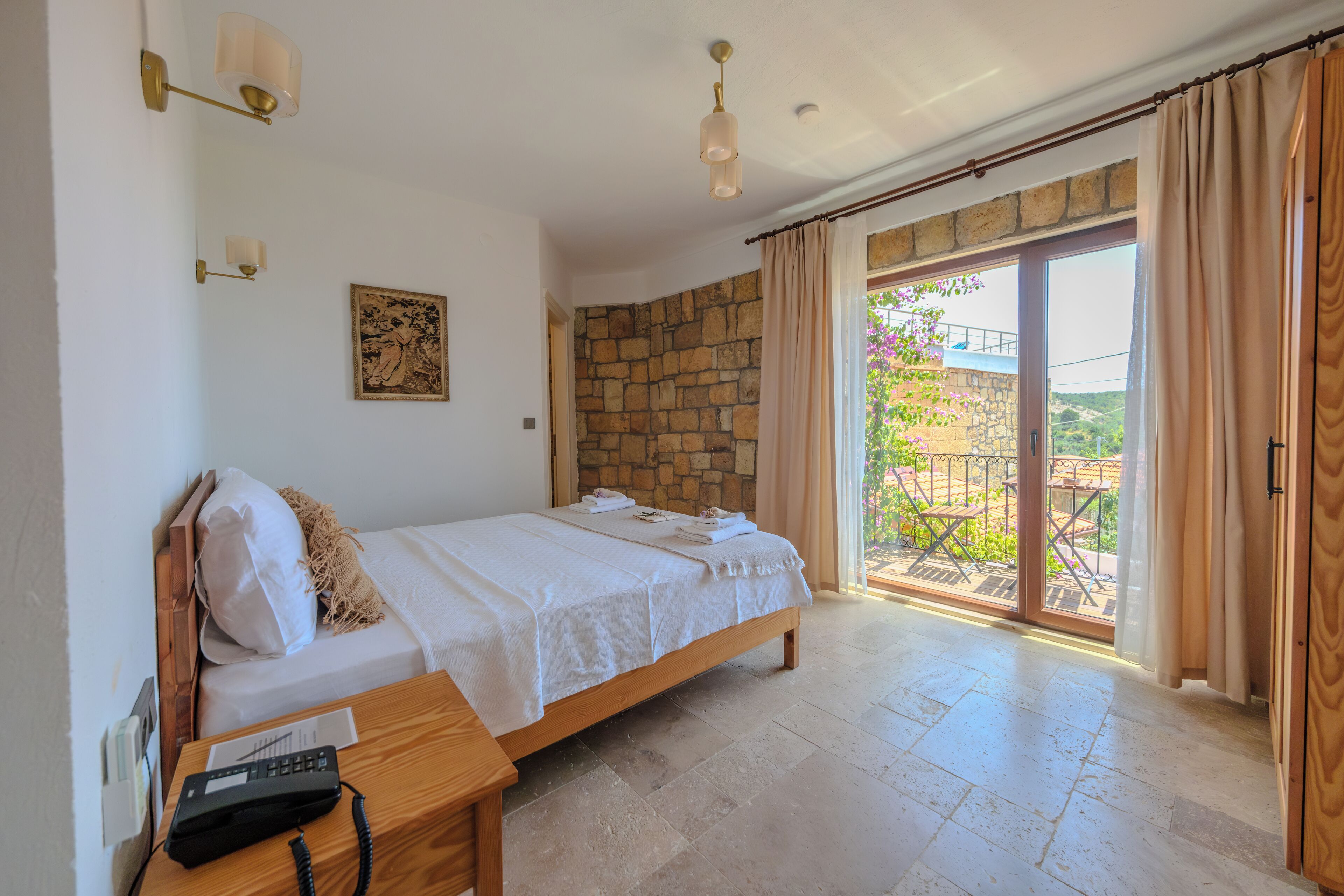 Un dormitorio con cama, mesita de noche de madera, teléfono, pared de piedra y vistas al exterior a través de una puerta de cristal.