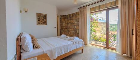 A bedroom with a bed, a wooden nightstand, a telephone, a stone wall, and a view of the outdoors through a glass door.