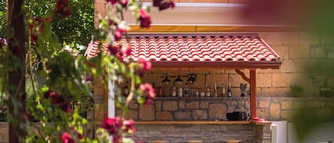A stone bar area with a tiled roof, surrounded by greenery and a stone wall.