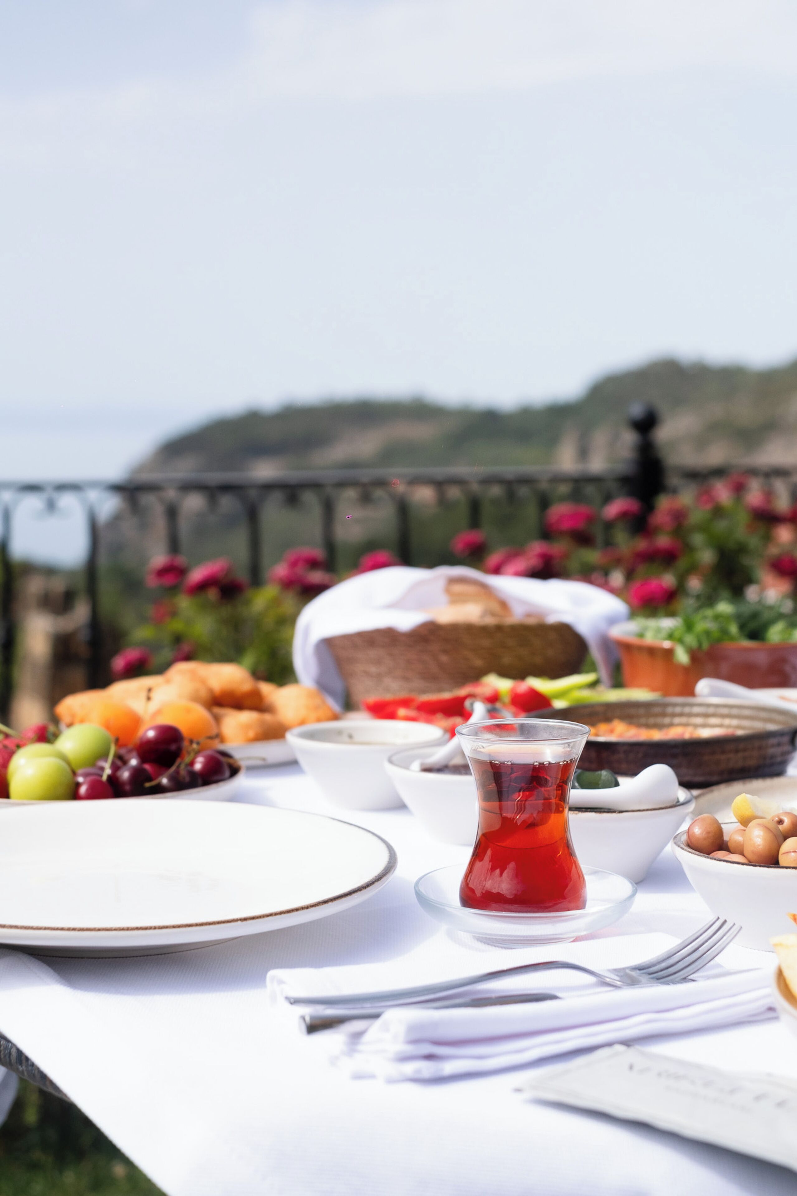 Una mesa servida con comida y bebidas, vista al mar y flores de fondo.