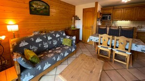 Een gezellige woonkamer met een bank, een houten salontafel en een eettafel met stoelen. Er is een keuken met houten kastjes en een magnetron.