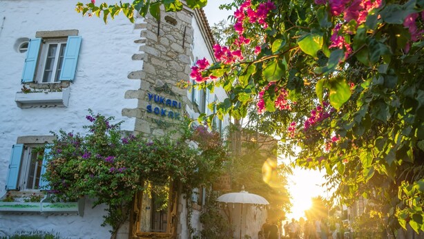 Een straat met keien, terrasjes, een stenen gebouw met blauwe luiken en bougainvillea bloemen.