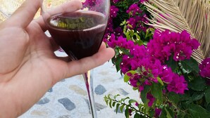 Een hand die een wijnglas vasthoudt, met in de achtergrond een stenen gebouw en kleurrijke bougainvillea's.